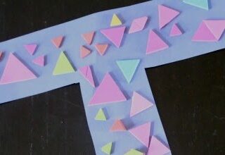 A blue paper cut into a T-shape is decorated with various colorful paper triangles in shades of pink, orange, green, and blue, arranged randomly across its surface. The T-shape is set against a dark background.