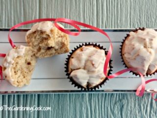 A rectangular tray holds three frosted muffins. One muffin is halved to show its inside. Red ribbon is artfully draped around the muffins. The tray is set on a textured, light-colored surface.
