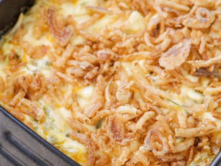 A casserole dish filled with a golden-brown, baked green bean casserole topped with crispy fried onions. Its placed on a checkered green cloth with a wooden-handled dish on the side.