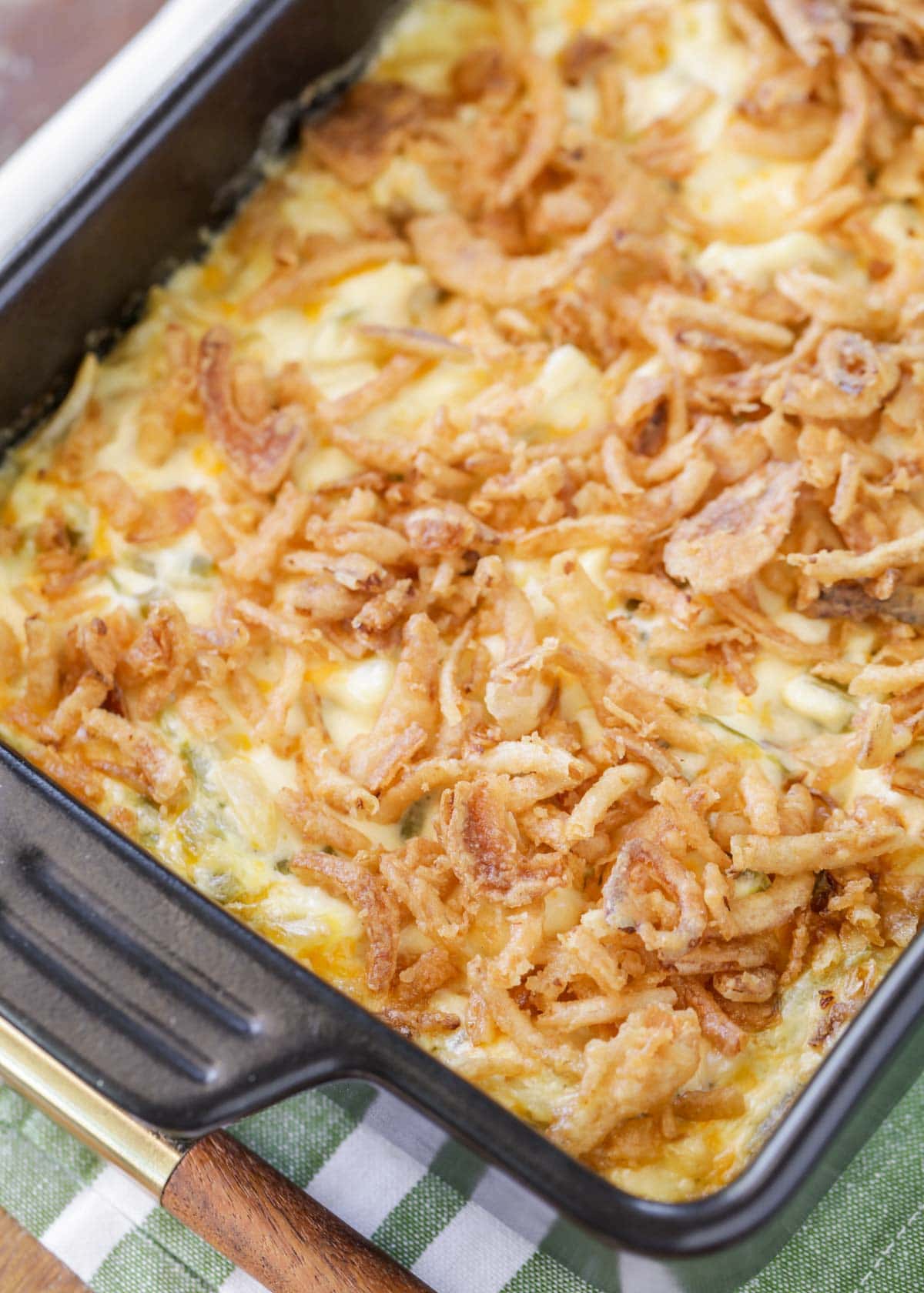A casserole dish filled with a golden-brown, baked green bean casserole topped with crispy fried onions. Its placed on a checkered green cloth with a wooden-handled dish on the side.