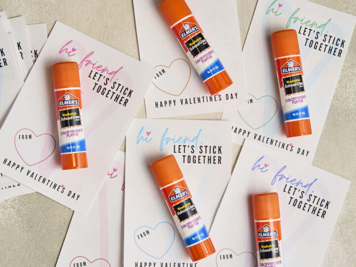 Multiple Elmers glue sticks are placed on Valentines cards with the message hi friend, lets stick together. Each card features a heart design and space for a from name. The background is a light, textured surface.