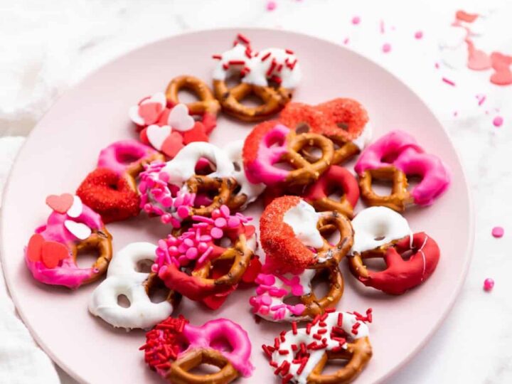 A pink plate with assorted pretzels covered in pink, red, and white candy coatings and sprinkles, some shaped like hearts, is on a white surface. Scattered sprinkles are visible around the plate.