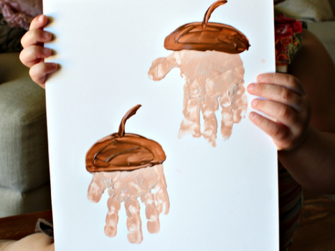 A child holds a painting showing two acorns, creatively made using brown paint and handprints for the acorns tops and beige paint for the bodies. The child is partially visible, smiling behind the artwork.