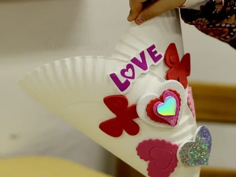 A childs hand holds a delightful paper craft cone adorned with red butterflies and pink glittery hearts, perfectly capturing the essence of Valentines Day. The word LOVE glimmers in purple letters, highlighting the charm and creativity of heartfelt paper crafts.