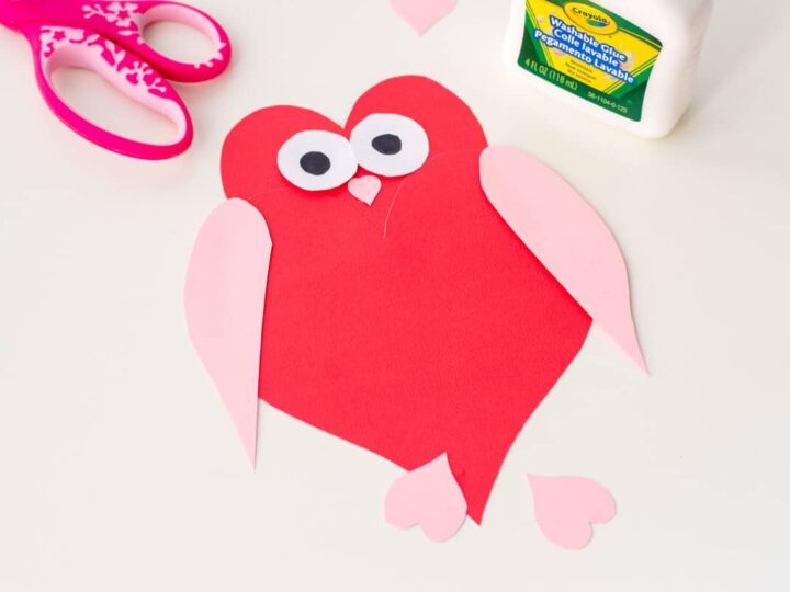 Crafted red heart-shaped owl with googly eyes, pink wings, and feet made of paper. Colorful scissors and a glue bottle are nearby on a white surface.