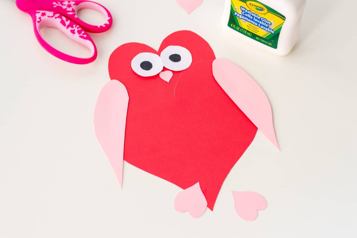Crafted red heart-shaped owl with googly eyes, pink wings, and feet made of paper. Colorful scissors and a glue bottle are nearby on a white surface.
