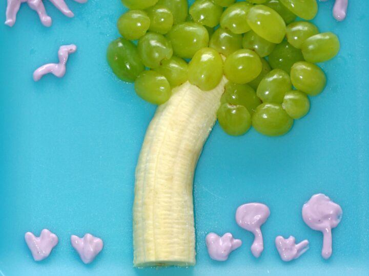 A creative snack on a blue plate resembling a tree: a banana as the trunk and green grapes as the foliage. Pink yogurt forms a sun, birds, and flowers. Text overlay reads, letter of the week snacks: T is for Tree.