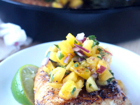 A grilled fish fillet topped with a pineapple and red onion salsa, garnished with a lime wedge. The dish is served on a white plate, and a black skillet with more food and a lime wedge is in the background.