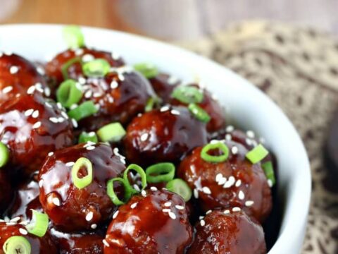 A bowl filled with glazed meatballs, topped with sesame seeds and sliced green onions. The meatballs appear coated in a glossy, dark sauce. A textured napkin is partially visible underneath the bowl.