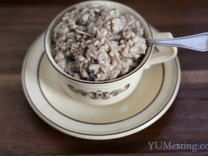 A beige ceramic teacup filled with oatmeal, placed on a matching saucer. A spoon rests inside the cup. The oatmeal appears to be cooked, with a creamy texture. The cup and saucer are decorated with a brown floral design.