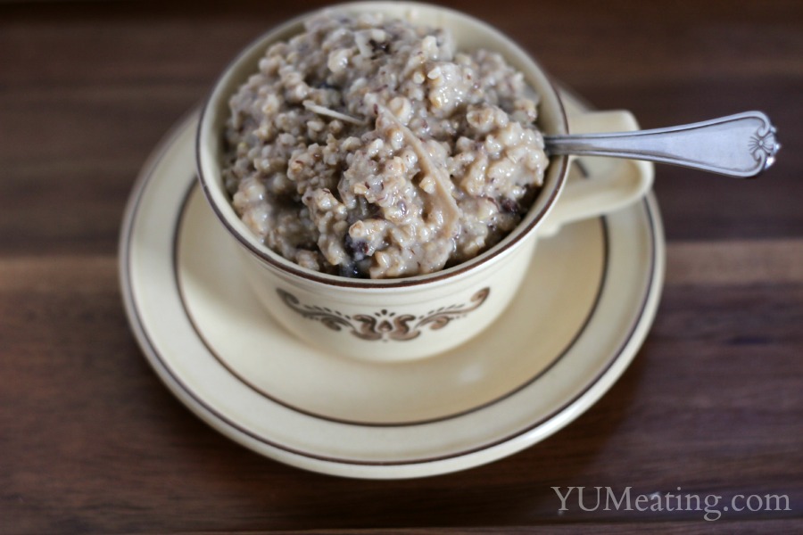 A beige ceramic teacup filled with oatmeal, placed on a matching saucer. A spoon rests inside the cup. The oatmeal appears to be cooked, with a creamy texture. The cup and saucer are decorated with a brown floral design.