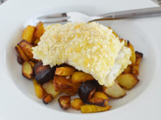 A white plate holds a serving of baked fish topped with breadcrumbs, resting on a bed of roasted potatoes and vegetables. A fork and knife are placed on the plates edge. The dish sits on a speckled countertop.