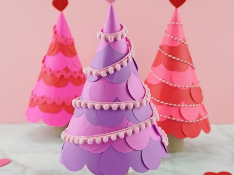 Three Valentines Day-themed trees made of colorful paper are displayed, with pink, red, and purple layers. Each tree is topped with a heart and decorated with string and paper scallops. They stand on a marble surface with small hearts scattered around.
