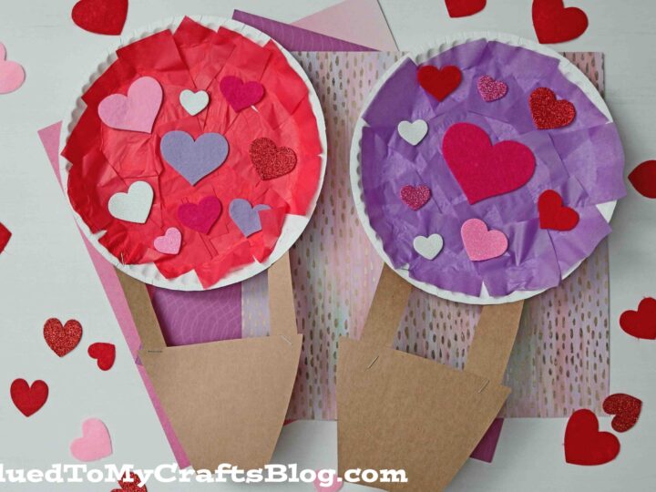 Two paper plate hot air balloons made from red and purple tissue paper and decorated with various heart-shaped cutouts. The baskets are made of brown paper. Surrounded by scattered decorative hearts on a pink background.