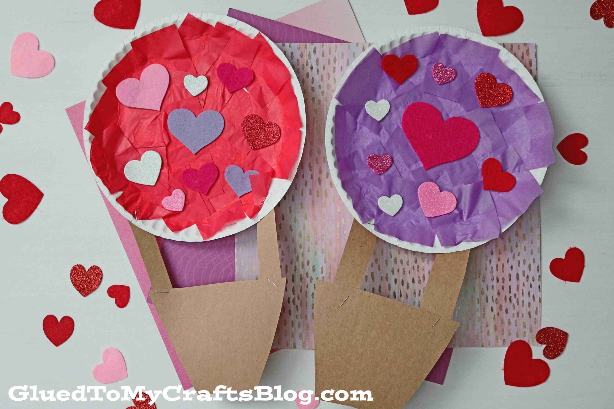 Two paper plate hot air balloons made from red and purple tissue paper and decorated with various heart-shaped cutouts. The baskets are made of brown paper. Surrounded by scattered decorative hearts on a pink background.