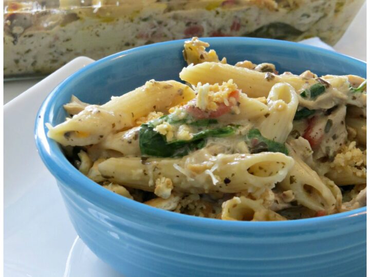 Close-up of a blue bowl filled with pesto chicken casserole, featuring penne pasta, spinach, and a creamy sauce, topped with breadcrumbs. A larger portion is in a glass baking dish in the background. Text reads Pesto Chicken Casserole at the bottom.