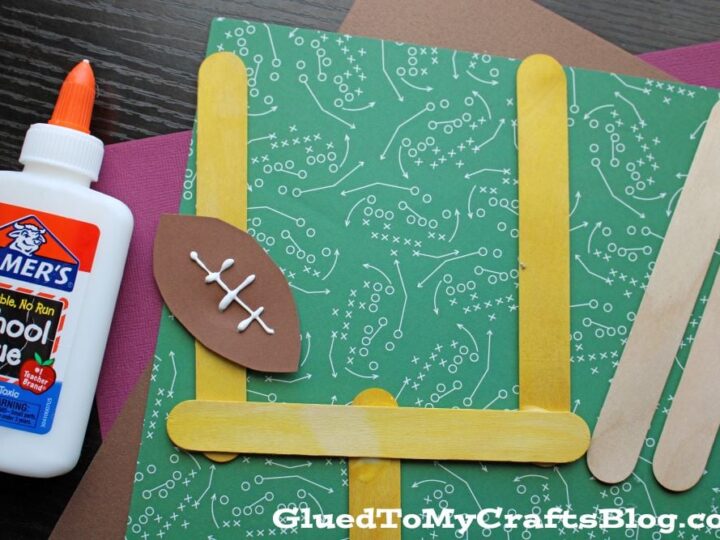 Craft project featuring a miniature football field made with popsicle sticks on patterned green paper. A small paper football with laces is in front. A bottle of Elmers glue is on the side. Text reads GluedToMyCraftsBlog.com.