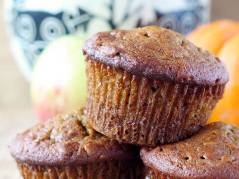 Three freshly baked muffins are stacked in the foreground, with a decorative bowl of more muffins and an apple in the background. The scene is set on a wooden surface, and a small pumpkin adds an autumnal touch.