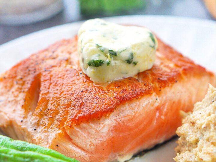 Grilled salmon topped with a dollop of herb butter on a white plate. Green beans are seen at the edge of the plate, alongside a serving of mashed potatoes. The salmon has a crispy, golden-brown crust.