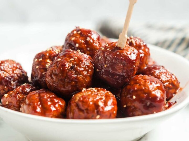 A white bowl filled with glazed meatballs, topped with sesame seeds. One meatball has a wooden toothpick inserted, ready to be picked up. The background is blurred, focusing on the dish.