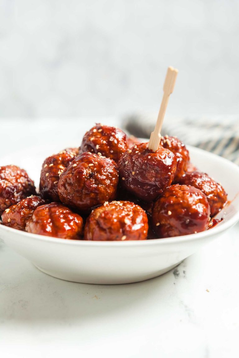 A white bowl filled with glazed meatballs, topped with sesame seeds. One meatball has a wooden toothpick inserted, ready to be picked up. The background is blurred, focusing on the dish.