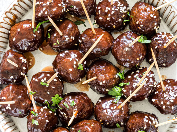 A plate filled with glazed meatballs garnished with sesame seeds and parsley, each with a toothpick. The meatballs are arranged neatly on a round, decorative plate.
