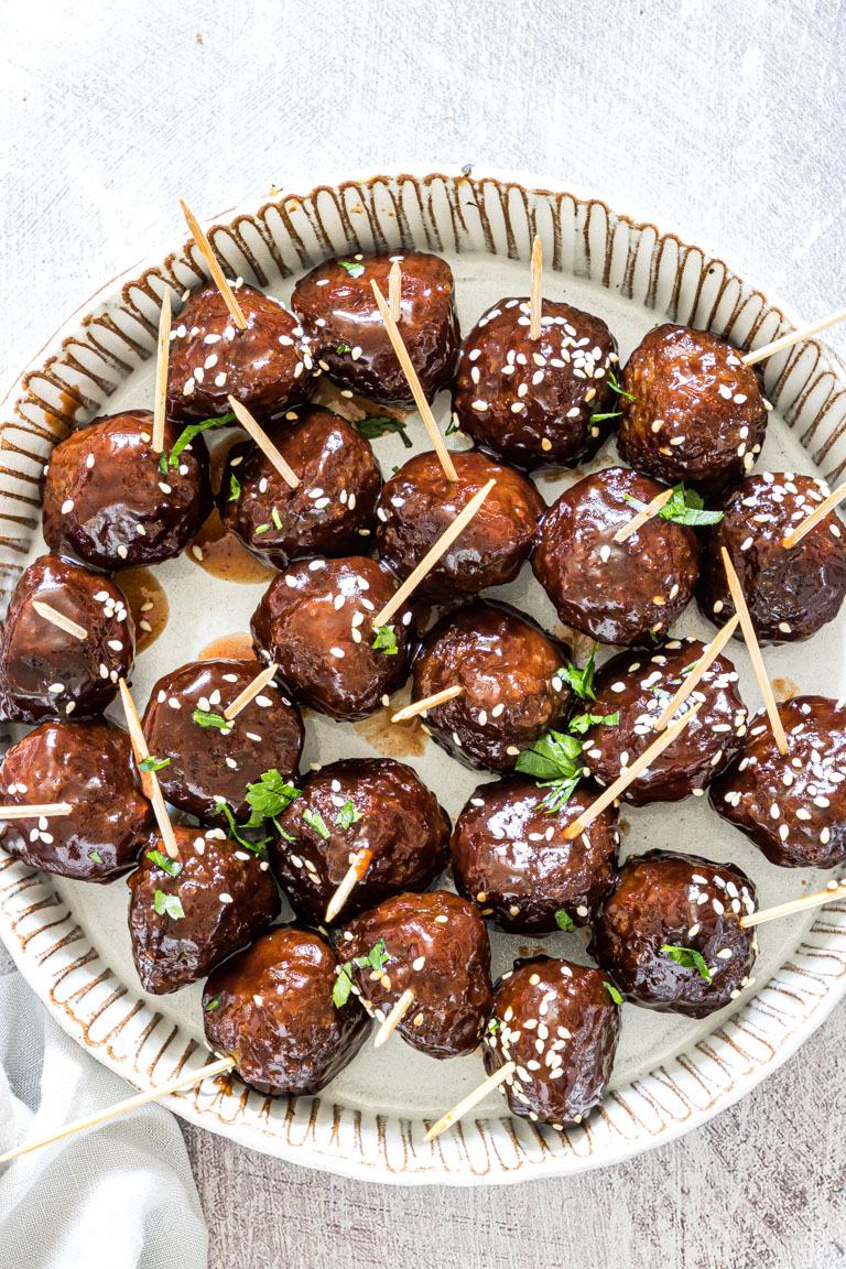 A plate filled with glazed meatballs garnished with sesame seeds and parsley, each with a toothpick. The meatballs are arranged neatly on a round, decorative plate.