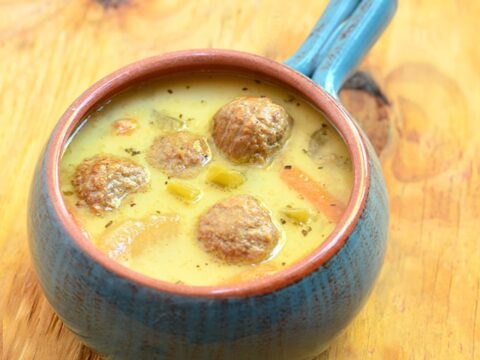 A blue ceramic bowl with a handle is filled with a creamy yellow soup, containing vegetables and meatballs. The bowl is placed on a wooden surface.