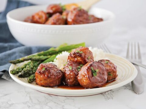 A plate of glazed meatballs garnished with herbs, served with white rice and roasted asparagus. A bowl of more meatballs is visible in the background. Silverware is placed beside the plate on a marble surface.
