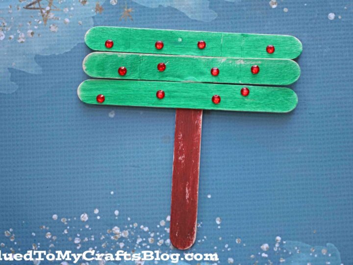 Craft of a Christmas tree made from green-painted popsicle sticks with red dots as ornaments, attached to a brown stick trunk, on a textured blue background with white speckles.