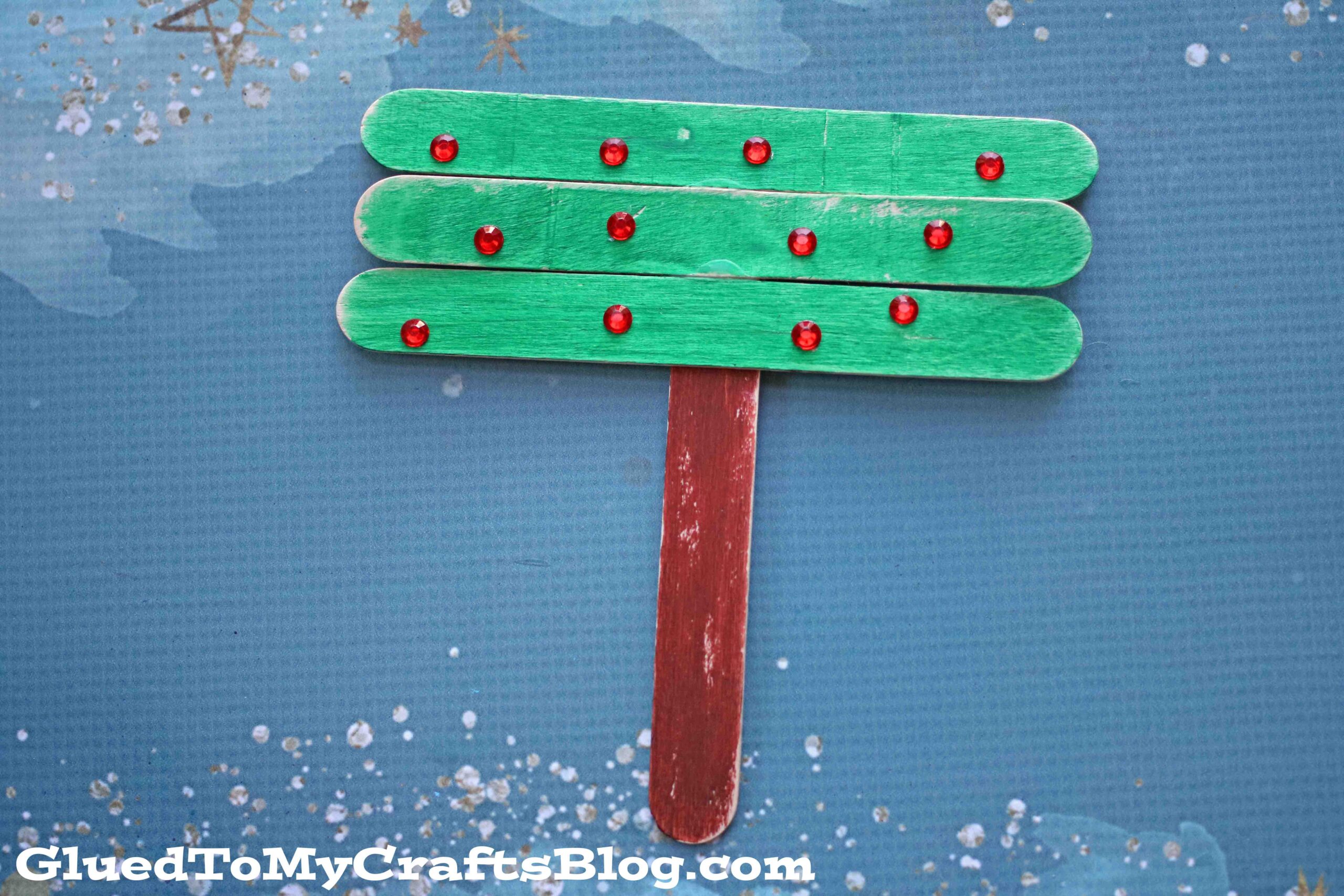 Craft of a Christmas tree made from green-painted popsicle sticks with red dots as ornaments, attached to a brown stick trunk, on a textured blue background with white speckles.