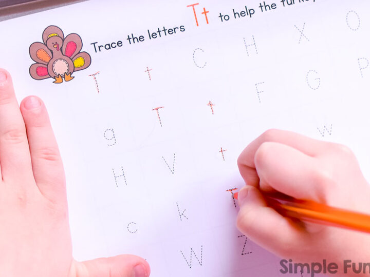 Child writing with an orange pencil on a worksheet featuring the letter T. The worksheet includes uppercase and lowercase letters with a cartoon turkey illustration. Text reads Trace the letters Tt to help the turkey.