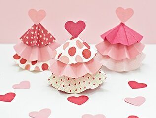 Three pink and red paper trees with heart toppers on a light pink background. The trees feature layered, ruffled patterns with polka dots and stripes. Scattered paper hearts decorate the surface around them.