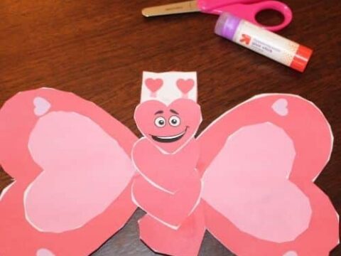 A butterfly-shaped craft made from red and pink paper with a happy face, decorated with heart patterns. It is placed on a wooden surface with a pair of scissors and a glue stick nearby.