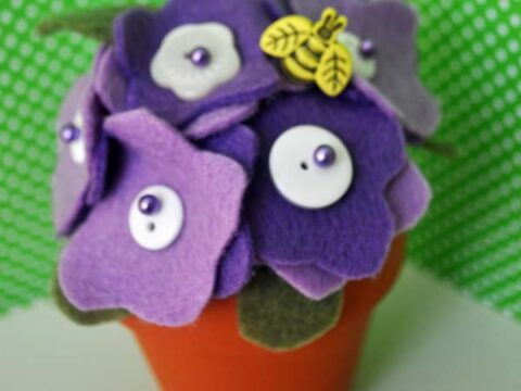 A small terracotta pot with purple and white felt flowers, adorned with buttons and pearl-like beads. A yellow charm with leaves is attached. Background features green fabric with white polka dots.