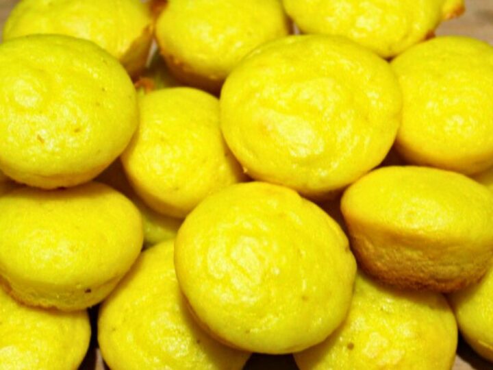 A pile of bright yellow muffins on a wooden surface. The muffins have smooth tops and are closely packed together.