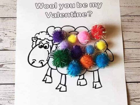 A coloring sheet with a cartoon sheep and the text Wool you be my Valentine? Colorful fluffy pom-poms are placed on the sheeps body, resembling wool. The background is a wooden surface.