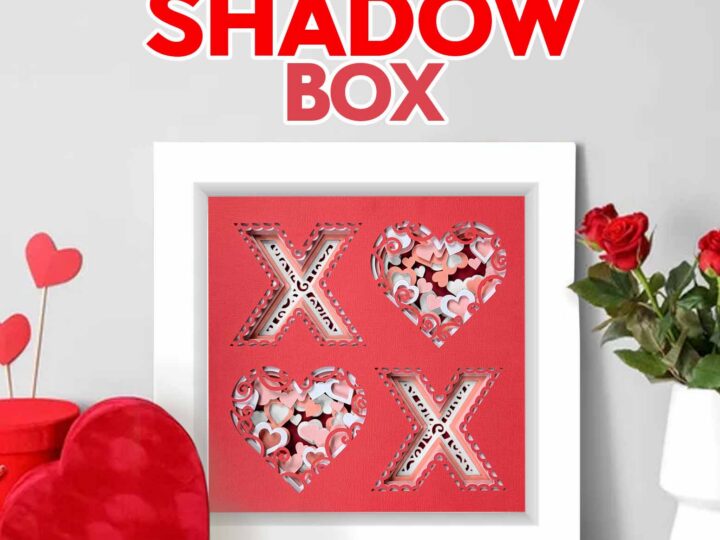 A framed love-themed shadow box with three cut-out shapes, two Xs and one heart, filled with red, pink, and white confetti. Its displayed next to red heart decorations and a vase of red roses on a white table.