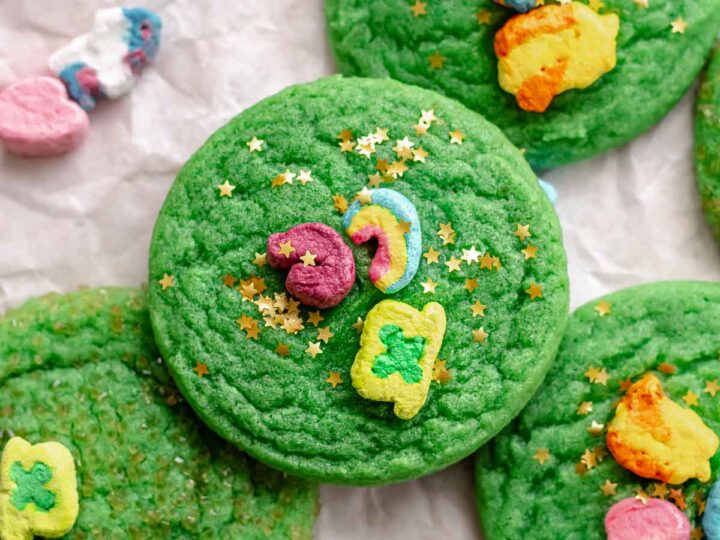 Bright green cookies topped with colorful marshmallow shapes and star sprinkles are scattered on a sheet of parchment paper. Some marshmallows are pink and rainbow-shaped, while others are yellow and shaped like shooting stars.