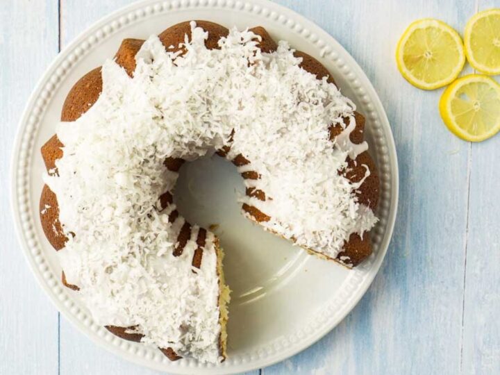 A Bundt cake on a white plate, topped with white frosting and shredded coconut, sits on a light wooden surface. Two slices have been removed, and three lemon slices are placed beside the plate.