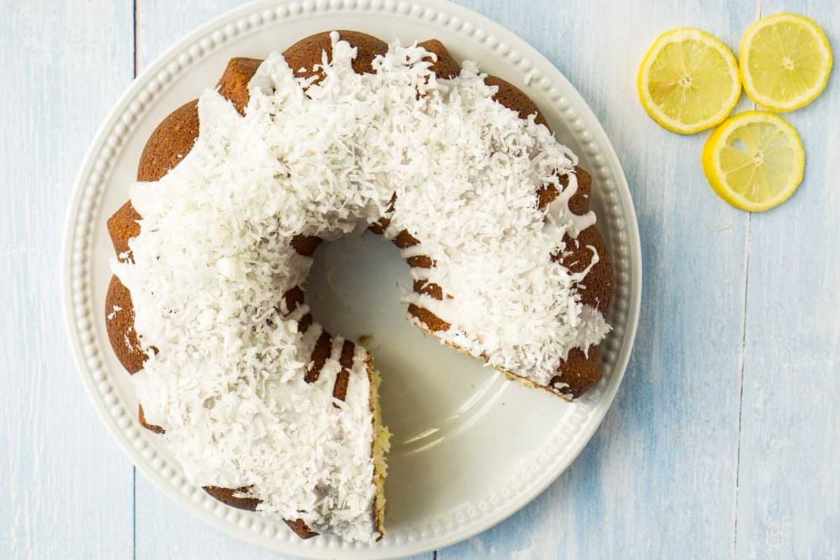 A Bundt cake on a white plate, topped with white frosting and shredded coconut, sits on a light wooden surface. Two slices have been removed, and three lemon slices are placed beside the plate.