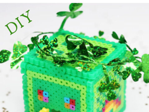 A colorful leprechaun trap made of Perler beads, designed as a small house with a rainbow door and shamrock decorations. The text reads Perler Bead Leprechaun Trap DIY with scattered gold confetti around.