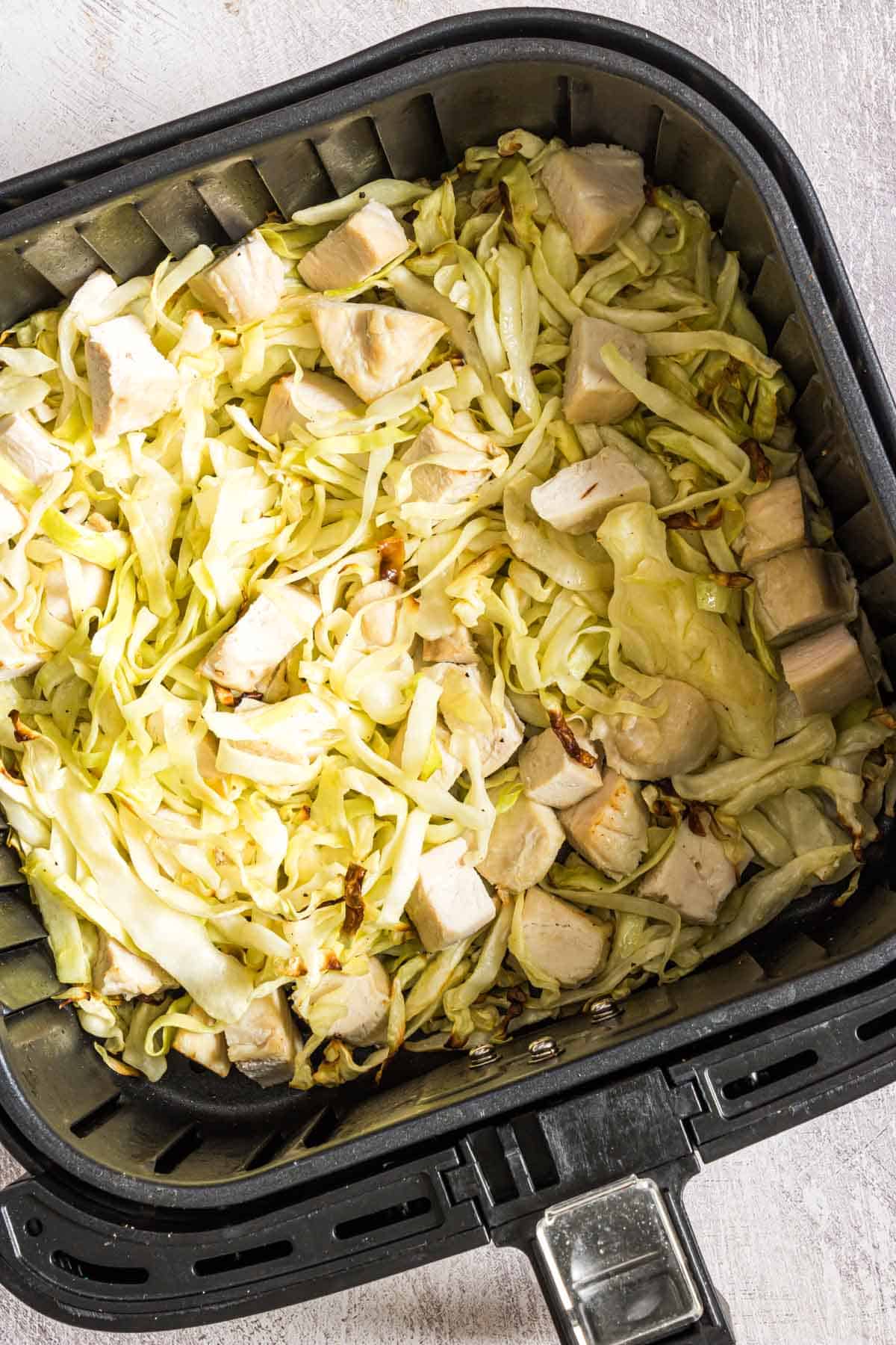 Chopped cabbage and diced chicken pieces in an air fryer basket, ready for cooking. The mixture is evenly spread, displaying a combination of pale green cabbage and lightly cooked white chicken chunks.