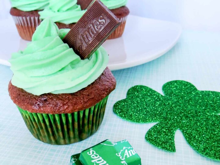 Three chocolate cupcakes topped with green frosting and Andes mint chocolate pieces. One cupcake is in the foreground, and two are on a white plate in the background. A glittery green shamrock and mint wrappers are next to the cupcakes.