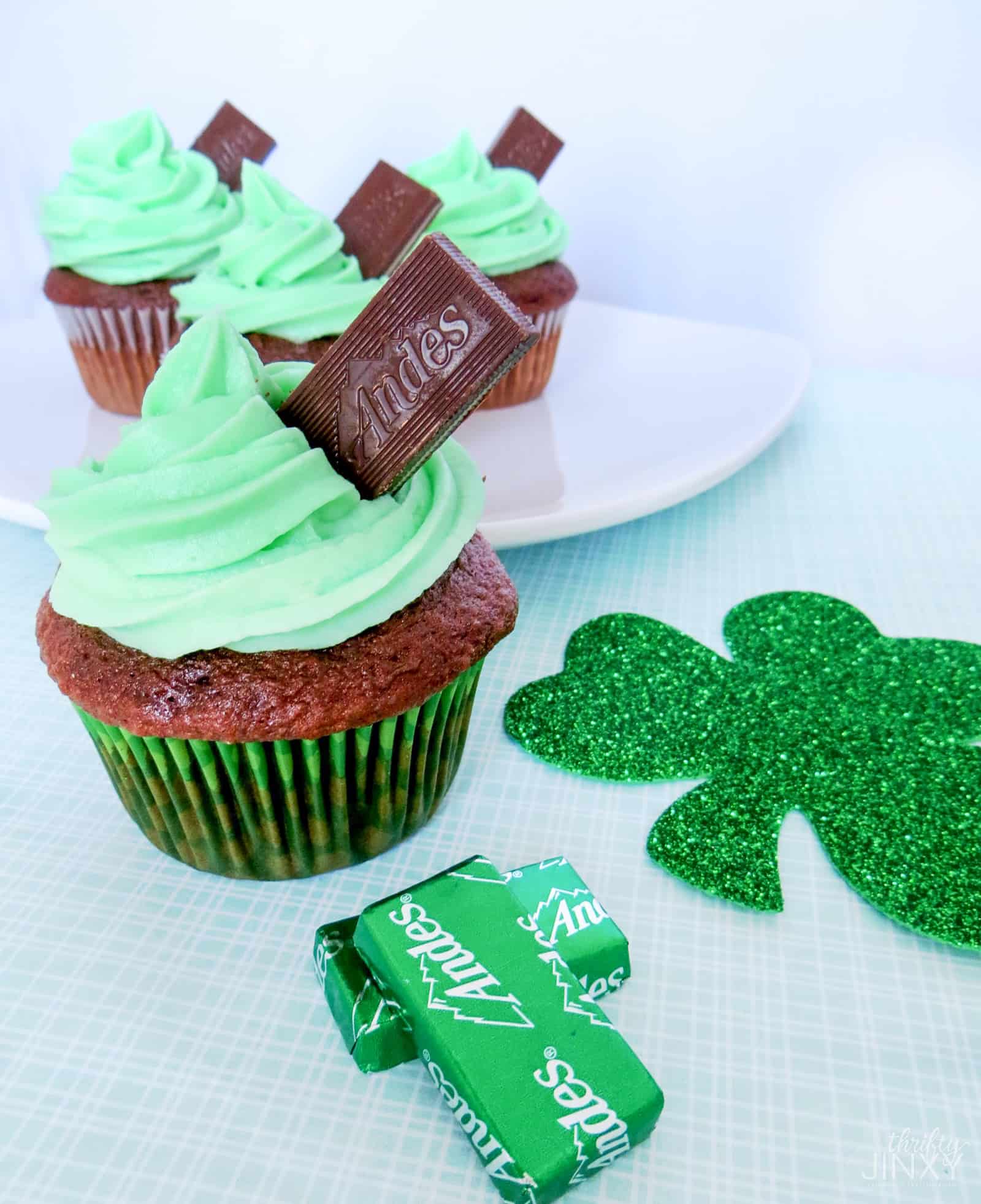 Three chocolate cupcakes topped with green frosting and Andes mint chocolate pieces. One cupcake is in the foreground, and two are on a white plate in the background. A glittery green shamrock and mint wrappers are next to the cupcakes.