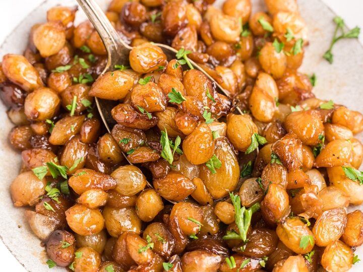 A close-up of glazed roasted grapes on a plate, garnished with fresh parsley. A spoon is picking up some of the grapes, and the dish appears glossy and caramelized.