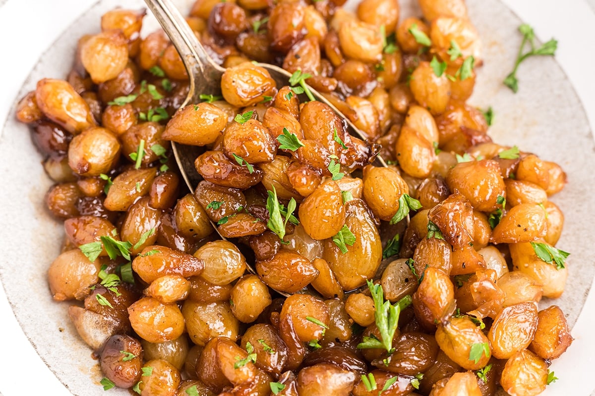 A close-up of glazed roasted grapes on a plate, garnished with fresh parsley. A spoon is picking up some of the grapes, and the dish appears glossy and caramelized.
