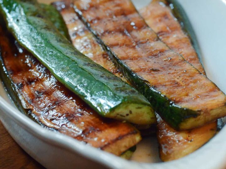 Grilled zucchini slices in a white, oval dish on a wooden surface. The zucchini has grill marks and a shiny, seasoned coating.