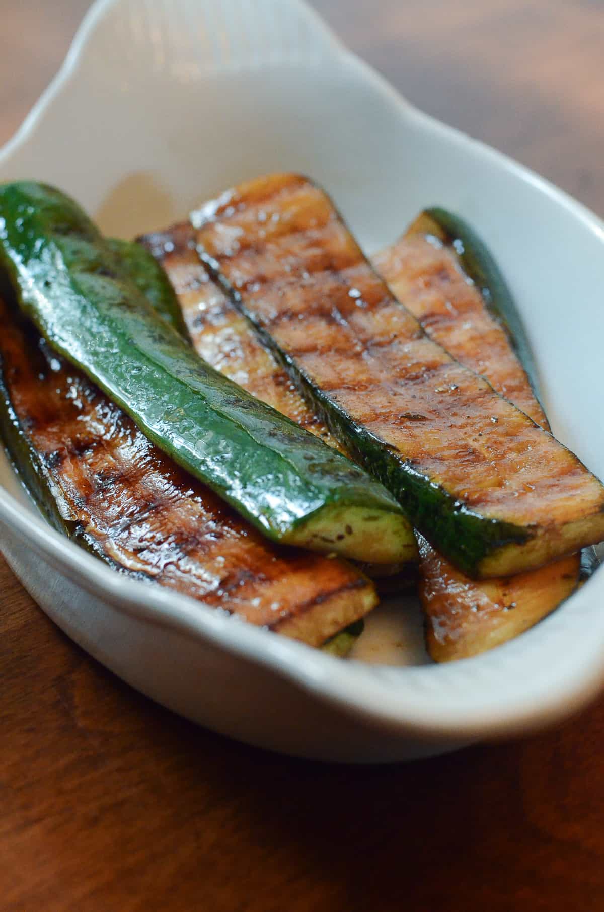 Grilled zucchini slices in a white, oval dish on a wooden surface. The zucchini has grill marks and a shiny, seasoned coating.