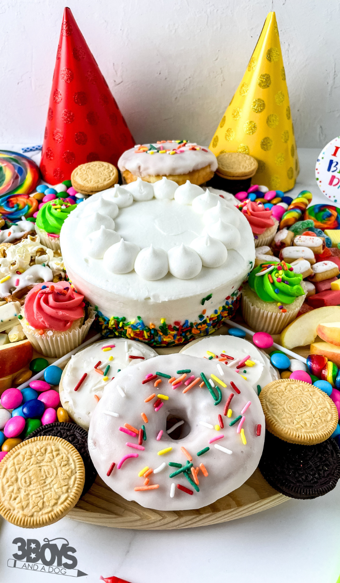A colorful birthday dessert board features a white-frosted cake at the center, surrounded by sprinkle-topped donuts, cookies, cupcakes, and assorted candies. Brightly colored party hats add to the festive vibe. Perfect for any party-themed occasion!.